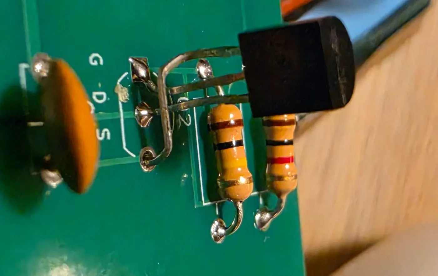 The 2N7000 on the circuit board with two of its legs bent and crossed over each other to reach the holes marked G and D, beside the silkscreen letters G, D and S